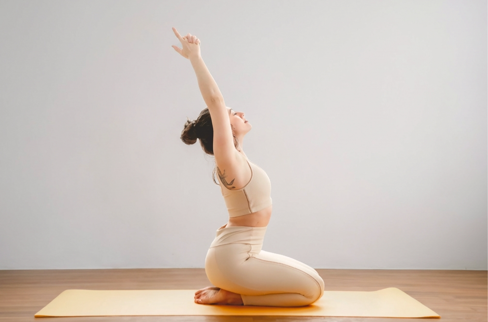 Женщина в бежевом топе и леггинсах выполняет йога-асану на коленях с руками вверх на жёлтом мате в студии